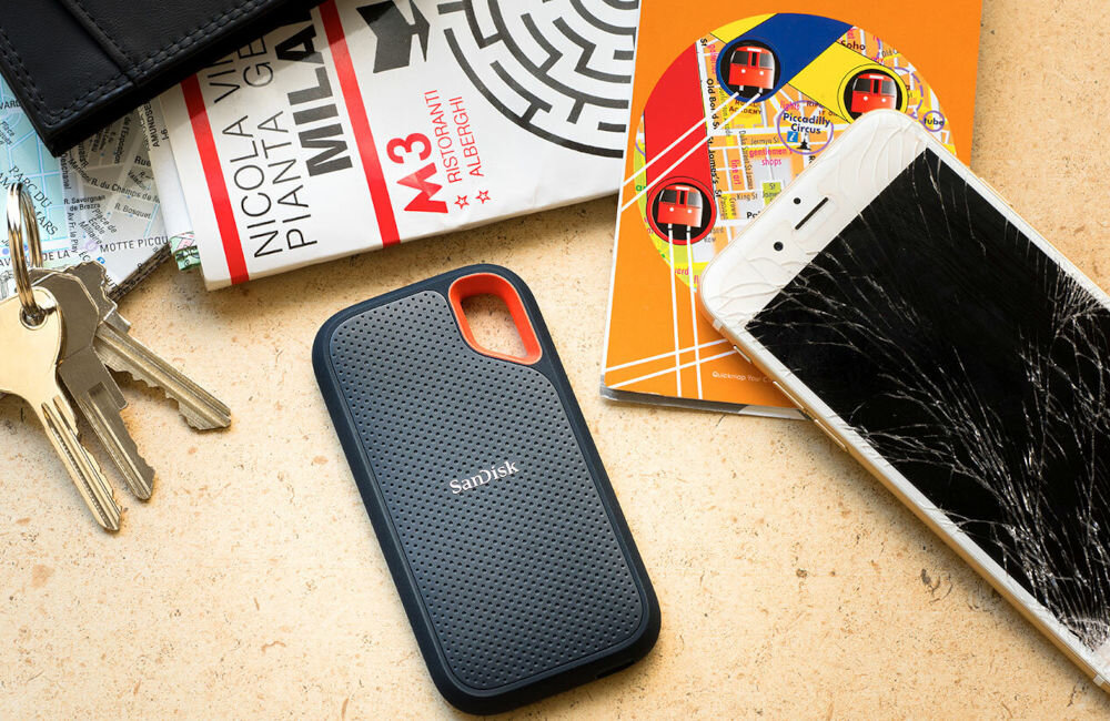 Na blacie leży przenośny dysk SSD marki SanDisk z napisem \'SanDisk\' na obudowie. Obok znajdują się klucze, przewodnik po Mediolanie oraz telefon z pękniętym ekranem.