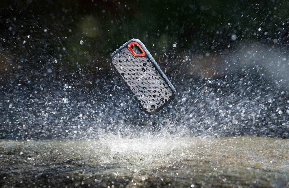 Na obrazie widać dysk przenośny w kształcie prostokąta z zaokrąglonymi rogami, unoszący się nad powierzchnią wody, z której rozpryskują się krople. Dysk ma ciemną obudowę z pomarańczowym akcentem i jest pokryty kroplami wody.