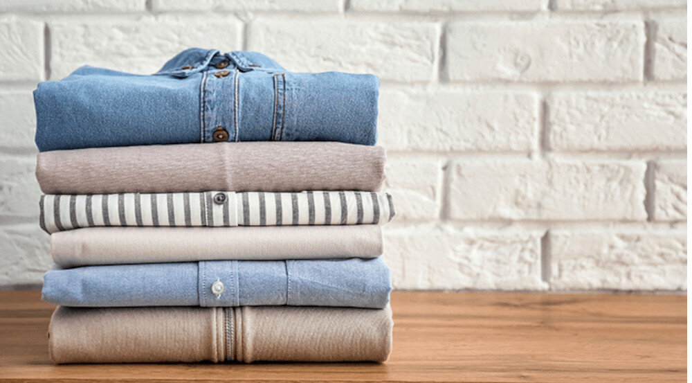 Stos złożonych ubrań leży na drewnianej powierzchni przed białą, ceglaną ścianą. Ubrania mają różne kolory i wzory, w tym dżinsowy, beżowy i w paski.