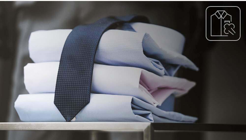 Na obrazie widoczne są cztery złożone koszule w jasnych kolorach, na których leży ciemny krawat. W prawym górnym rogu znajduje się ikona przedstawiająca koszulę i symbol pary.