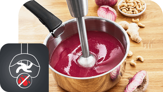 Na drewnianym blacie stoi metalowy garnek z czerwonym kremem, w którym zanurzony jest blender ręczny. Obok garnka leżą kawałki cebuli, orzechy nerkowca i imbir, a w lewym dolnym rogu widoczna jest ikona z przekreślonym symbolem noża.