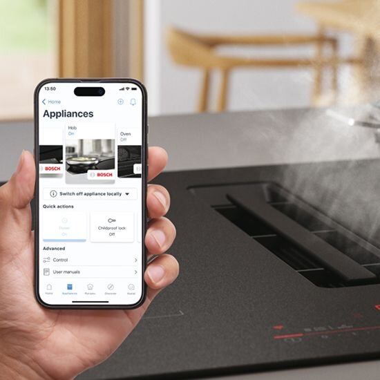 Osoba trzyma smartfon z aplikacją \'Appliances\', na ekranie widoczne są opcje sterowania urządzeniami marki BOSCH. Obok znajduje się płyta indukcyjna z wbudowanym okapem, z której unosi się para.