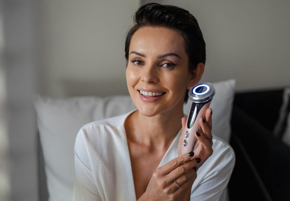 Osoba trzyma urządzenie do mezoterapii bezigłowej z podświetlanym elementem. W tle widoczne są poduszki na kanapie.