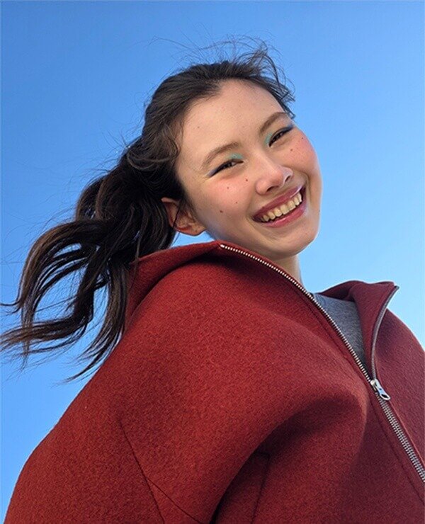 Osoba w czerwonej bluzie z kapturem uśmiecha się na tle błękitnego nieba.