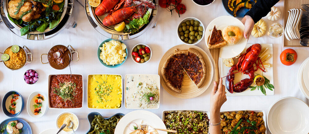 Stół zastawiony różnorodnymi potrawami, w tym homarami, pieczonym mięsem, sałatkami i deserami. Na stole znajdują się również miski z oliwkami, sosami i pieczywem.