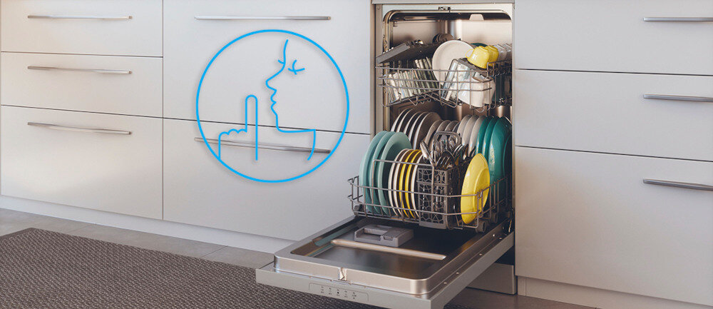Otwarta zmywarka z widocznymi naczyniami w środku, umieszczona w kuchennej zabudowie. Obok zmywarki znajduje się niebieski symbol przedstawiający twarz z palcem przy ustach, sugerujący cichą pracę urządzenia.