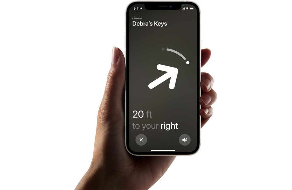 Osoba trzyma smartfon z aplikacją pokazującą lokalizację kluczy. Na ekranie widoczny jest duży biały symbol strzałki oraz tekst: \'Debra\'s Keys\' i \'20 ft to your right\'.