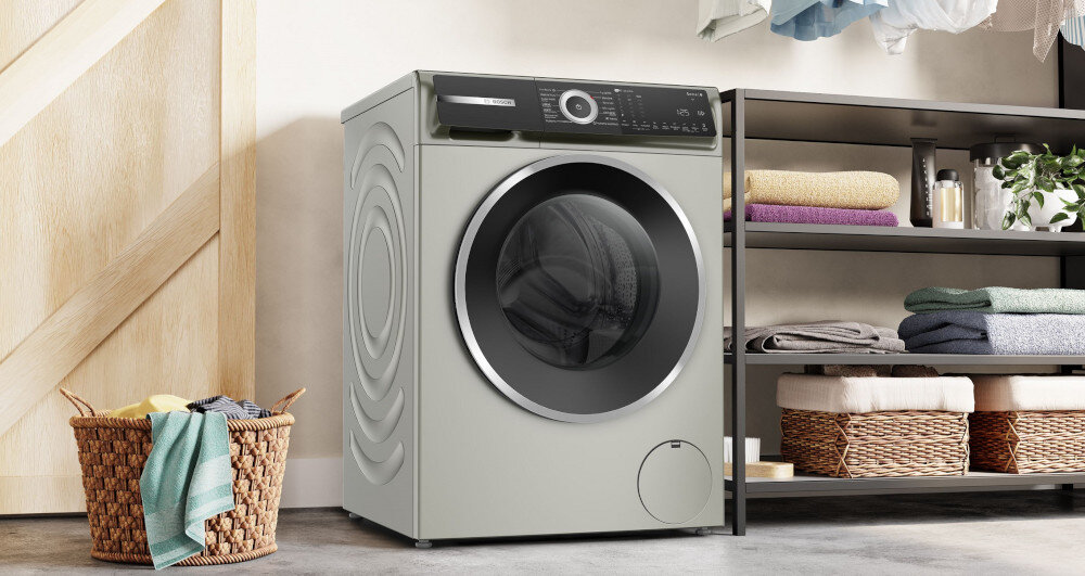 Pralka w kolorze srebrnym z dużym, okrągłym oknem na drzwiach, stoi obok półki z ręcznikami i koszem na pranie. Na panelu sterowania widoczne są przyciski i pokrętło.