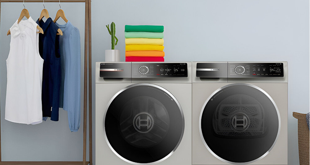 Na obrazie widoczne są dwie pralki marki Bosch z czarnymi drzwiczkami i panelami sterowania. Obok pralek znajduje się wieszak z trzema koszulami oraz stos kolorowych ręczników na pralce.