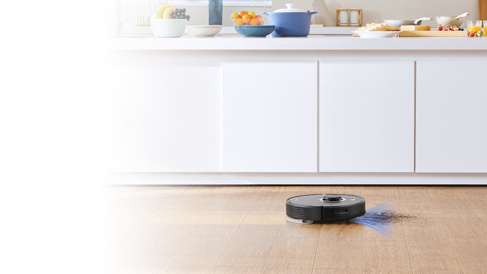Robot sprzątający na drewnianej podłodze w kuchni z białymi szafkami. Na blacie znajdują się różne naczynia i owoce.