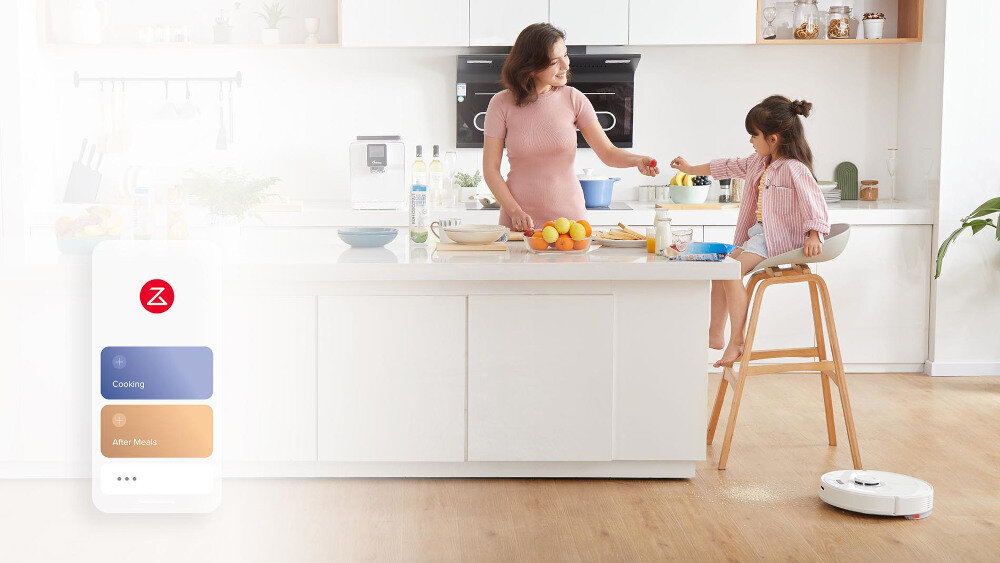 W kuchni znajduje się wyspa kuchenna z owocami i naczyniami. Na podłodze widoczny jest biały robot sprzątający, a na ekranie telefonu widnieje logo z literą Z oraz teksty \'Cooking\' i \'After Meals\'.