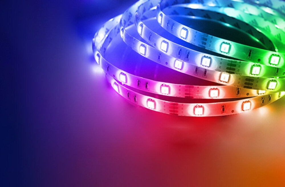Na obrazie widoczna jest taśma LED emitująca światło w różnych kolorach, takich jak niebieski, różowy, czerwony i zielony. Taśma jest zwinięta w kilka pętli na jednolitym tle.