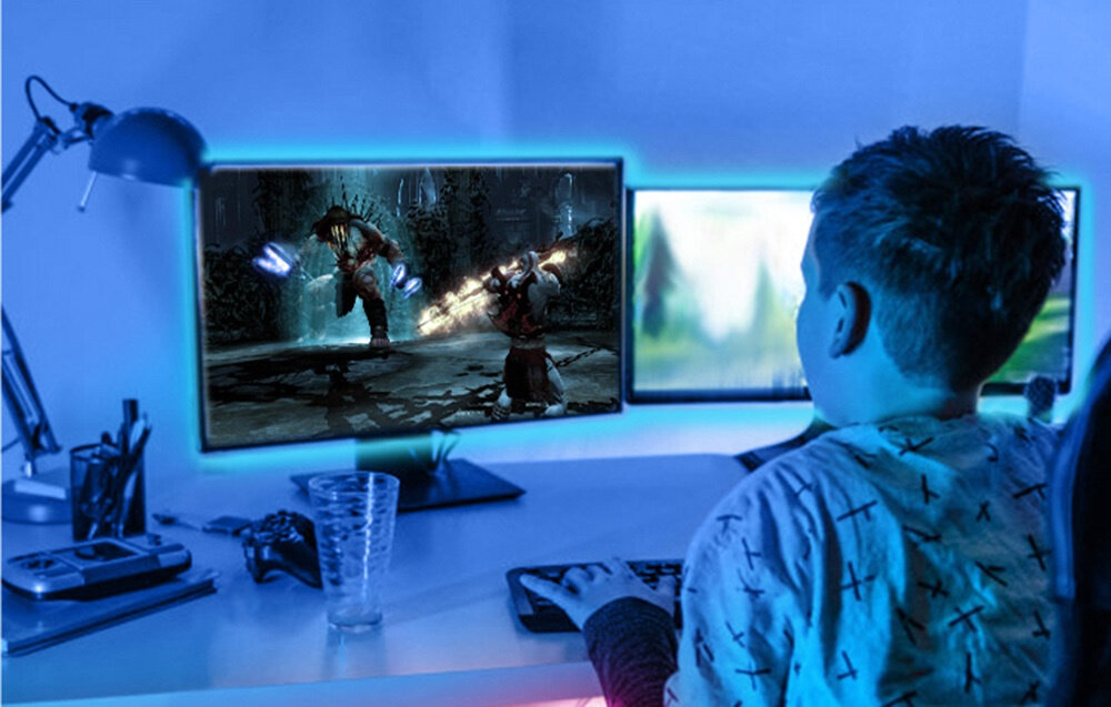 Osoba siedzi przy biurku z dwoma monitorami, z których jeden wyświetla grę wideo. Wokół monitorów widoczna jest niebieska poświata, prawdopodobnie pochodząca od taśmy LED.