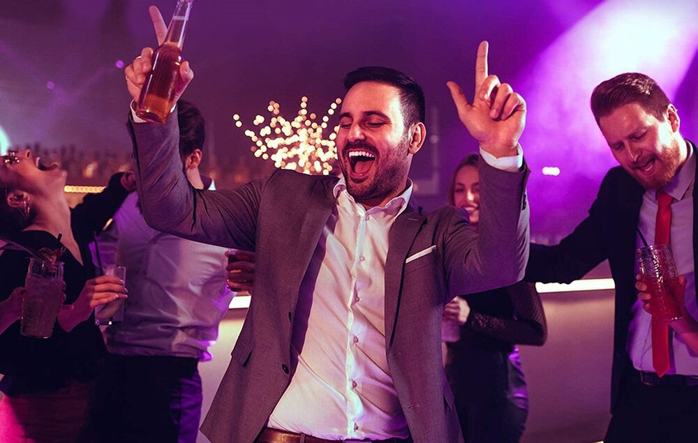 Grupa ludzi bawi się na imprezie, trzymając napoje w rękach. W tle widoczne są kolorowe światła i dekoracje.