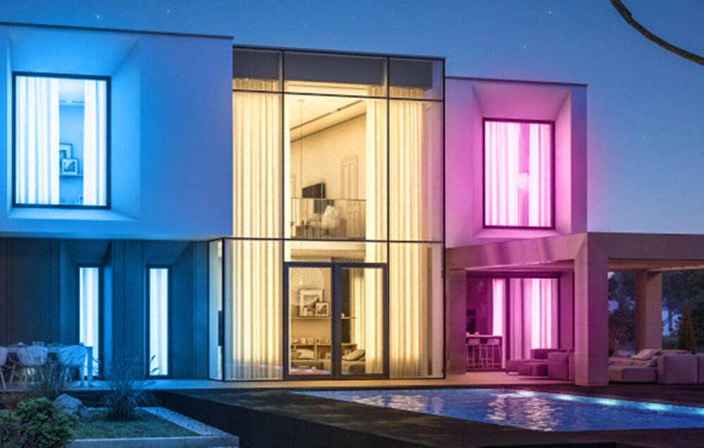 Nowoczesny dom z dużymi oknami, oświetlony różnokolorowymi światłami LED: niebieskim, żółtym i różowym. Przed budynkiem znajduje się basen, a wnętrze jest widoczne przez przeszklone ściany.