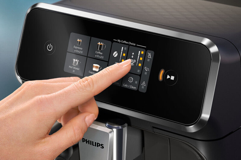 Osoba dotyka panelu dotykowego ekspresu do kawy, na którym widoczne są opcje takie jak Espresso, Coffee, Hot Water i Cappuccino. Na obudowie ekspresu znajduje się logo PHILIPS.