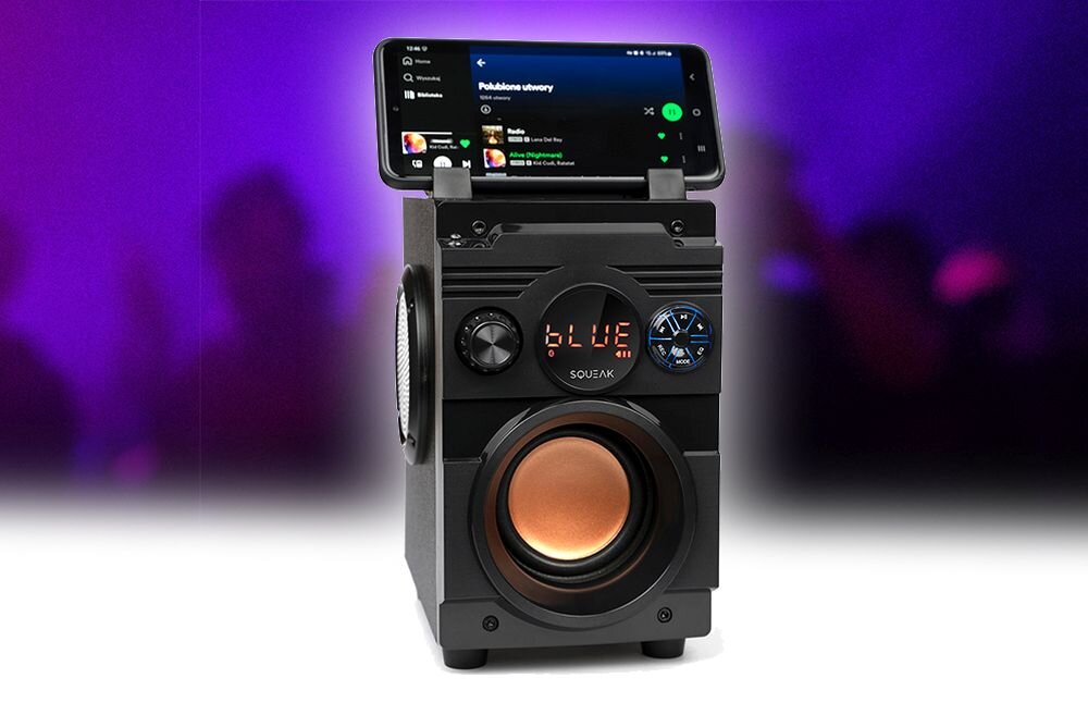 Czarny głośnik mobilny z dużym, miedzianym głośnikiem niskotonowym i wyświetlaczem LED pokazującym napis \'BLUE\'. Na górze urządzenia znajduje się smartfon z otwartą aplikacją muzyczną.