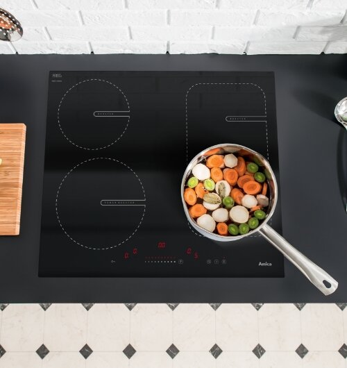 Czarna płyta gazowo-ceramiczna z czterema polami grzewczymi, na której stoi garnek z pokrojonymi warzywami. Widoczne logo \'Amica\' oraz panel dotykowy z czerwonymi oznaczeniami.