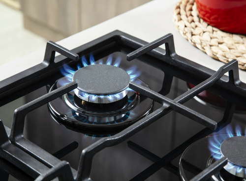 Na obrazie widoczna jest płyta gazowo-ceramiczna z palnikiem, na którym pali się niebieski płomień. W tle znajduje się czerwona pokrywka na plecionym podkładzie.