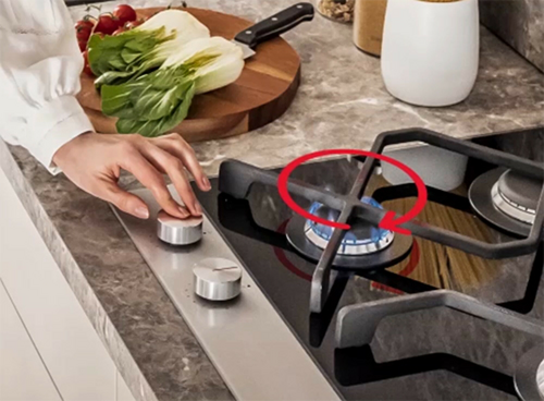 Osoba reguluje pokrętło na płycie gazowo-ceramicznej z płonącym palnikiem. Na blacie obok znajdują się warzywa i nóż na desce do krojenia.