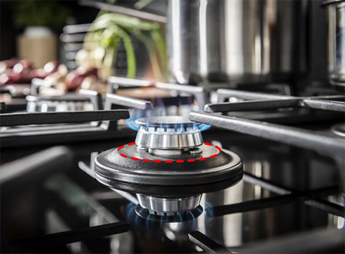 Na obrazie widoczny jest palnik gazowy z niebieskim płomieniem na płycie kuchennej. W tle znajdują się rozmyte elementy kuchenne, takie jak garnki i rośliny.