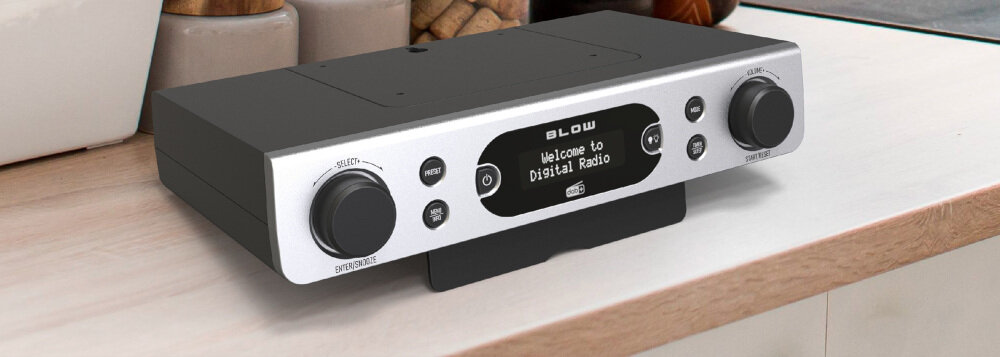 Srebrne przenośne radio z czarnym panelem i wyświetlaczem pokazującym tekst \'Welcome to Digital Radio\'. Na panelu znajdują się dwa pokrętła oraz kilka przycisków, a nad wyświetlaczem widoczne jest logo \'BLOW\'.