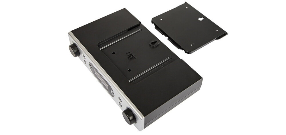 Na zdjęciu widoczny jest srebrny, przenośny radioodtwarzacz z czarnymi elementami i dwoma pokrętłami. Obok znajduje się czarna, prostokątna pokrywa z otworami montażowymi.