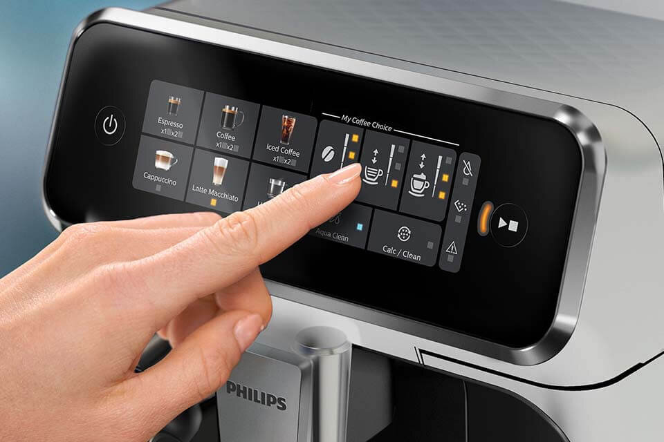 Osoba dotyka panelu dotykowego ekspresu do kawy, na którym widoczne są opcje wyboru napojów, takie jak Espresso, Coffee, Iced Coffee, Cappuccino i Latte Macchiato. Na obudowie ekspresu widnieje logo PHILIPS.