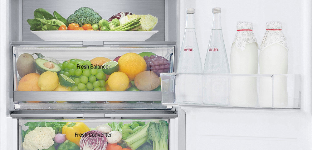 Wnętrze lodówki z widocznymi półkami wypełnionymi różnorodnymi warzywami i owocami. Na drzwiach znajdują się dwie butelki wody z etykietą \'evian\' oraz dwie butelki mleka.