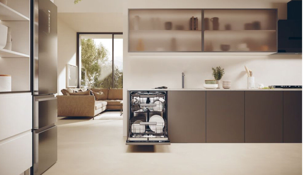 Kuchnia z otwartą zmywarką w zabudowie, wypełnioną naczyniami. W tle widoczny jest salon z sofą i dużym oknem.