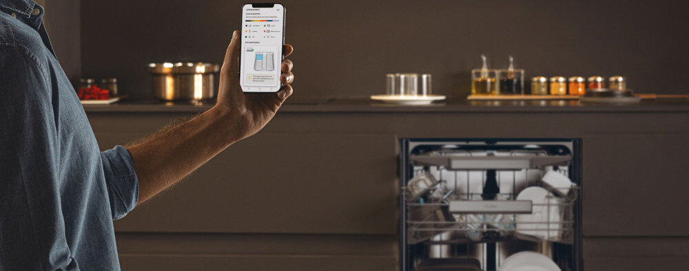 Osoba trzyma smartfon z aplikacją pokazującą ustawienia zmywarki. W tle otwarta zmywarka z widocznymi naczyniami w środku.