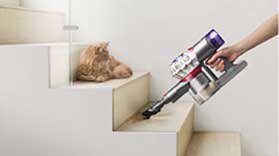 Osoba używa odkurzacza ręcznego do czyszczenia schodów, obok leży kot. Odkurzacz ma metaliczny korpus z fioletowymi i czerwonymi elementami.