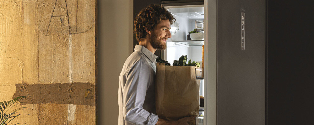 Osoba stoi przed otwartą lodówką, trzymając papierową torbę z zakupami. Wnętrze lodówki jest oświetlone i widać w nim kilka produktów spożywczych.