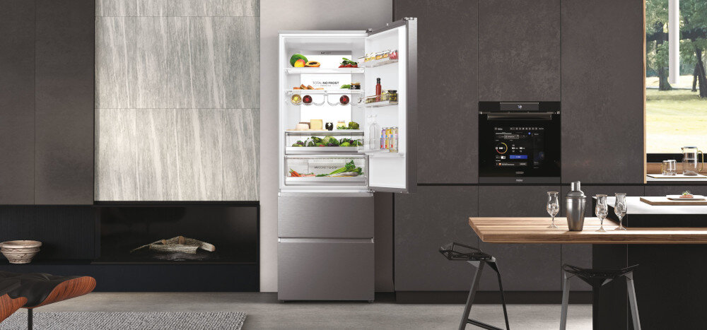 Kuchnia z otwartą lodówką w kolorze platynowy inox, wypełnioną różnorodnymi produktami spożywczymi. Obok znajduje się zabudowany piekarnik, a na stole stoją szklanki i karafka.