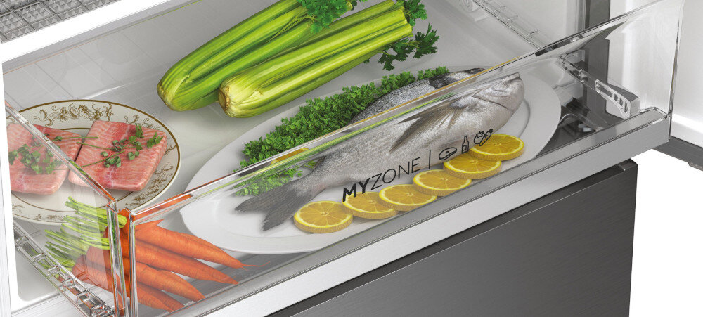 Wnętrze lodówki z przezroczystą szufladą oznaczoną napisem \'MYZONE\', w której znajdują się warzywa, talerz z rybą i plasterkami cytryny. Obok ryby leżą seler, marchewki i kawałki mięsa na ozdobnym talerzu.