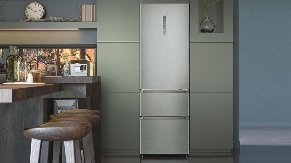 W kuchni znajduje się lodówka w kolorze ciemnego inoxu, wkomponowana w zabudowę meblową. Obok widoczny jest fragment blatu z krzesłami barowymi oraz różnymi przedmiotami kuchennymi.