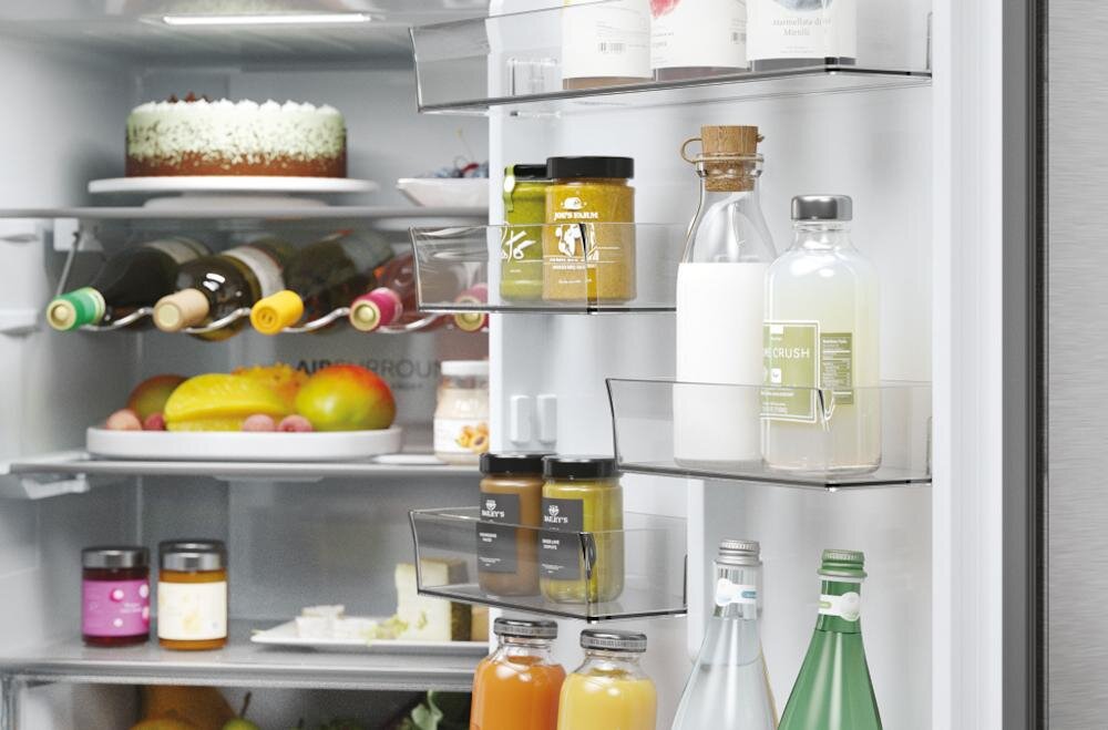 Wnętrze lodówki z widocznymi półkami wypełnionymi różnymi produktami spożywczymi, takimi jak butelki, słoiki i owoce. Na górnej półce znajduje się tort, a poniżej widoczne są butelki z winem oraz miska z owocami.