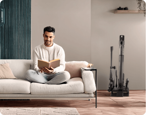 Wnętrze pokoju z sofą, na której siedzi osoba z książką. Obok stoi odkurzacz pionowy na stacji dokującej.