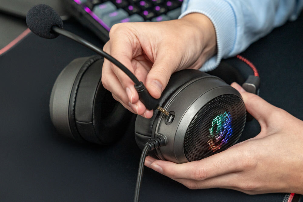 Osoba trzyma słuchawki nauszne z podłączanym mikrofonem. Na zewnętrznej stronie muszli widoczny jest kolorowy wzór świetlny.