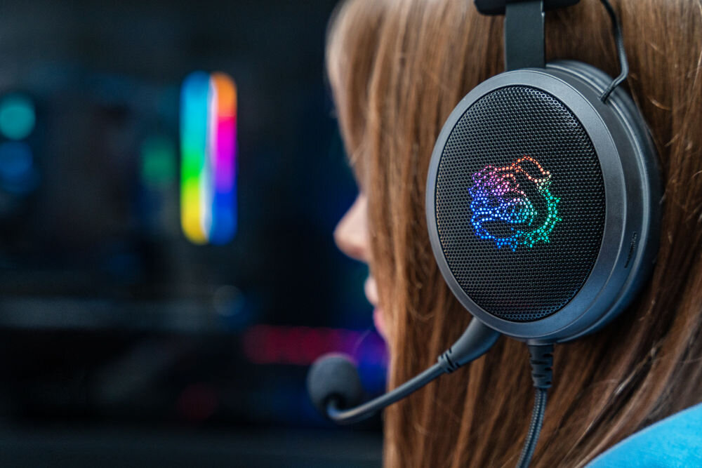 Osoba nosi nauszne słuchawki z widocznym kolorowym logo w kształcie psa. W tle widać rozmyte światła RGB.