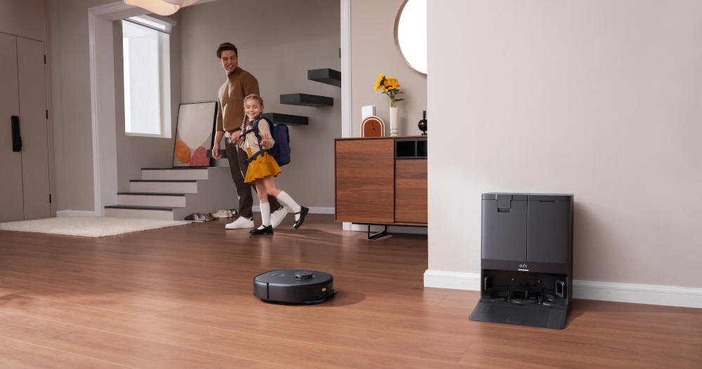 W przestronnym wnętrzu z drewnianą podłogą znajduje się robot sprzątający oraz stacja dokująca z logo \'eufy\'. W tle widać osobę dorosłą i dziecko, a także schody i komodę z dekoracjami.
