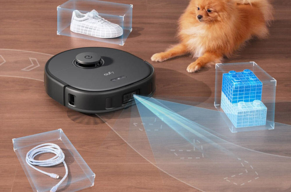 Na drewnianej podłodze znajduje się czarny robot sprzątający z napisem \'eufy\', emitujący niebieski promień. Wokół robota są przezroczyste pudełka z białym butem, niebieskimi klockami i białym kablem, a obok leży mały pies.