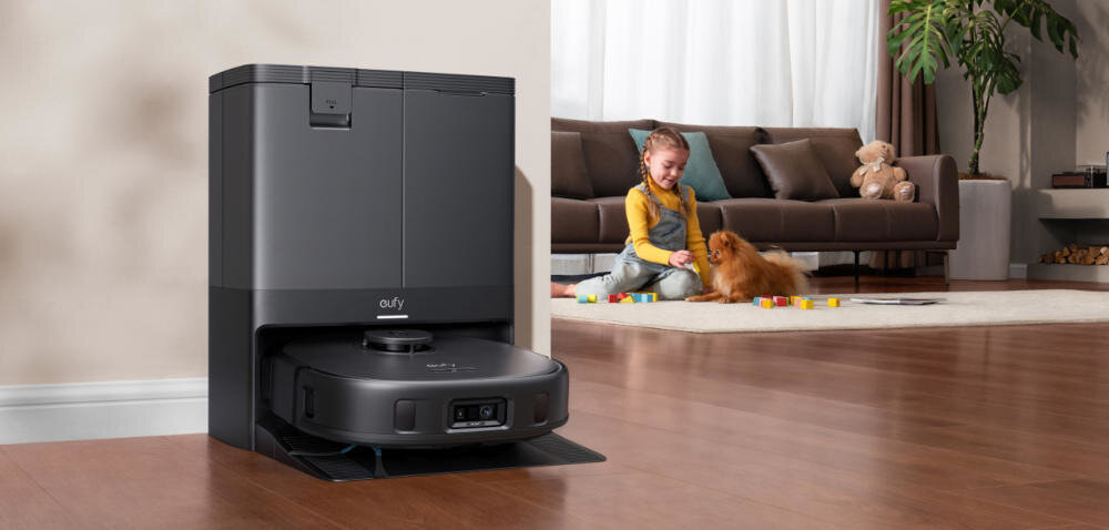 Na drewnianej podłodze stoi czarny robot sprzątający z widocznym logo \'eufy\'. W tle dziecko bawi się z psem na dywanie obok kanapy.