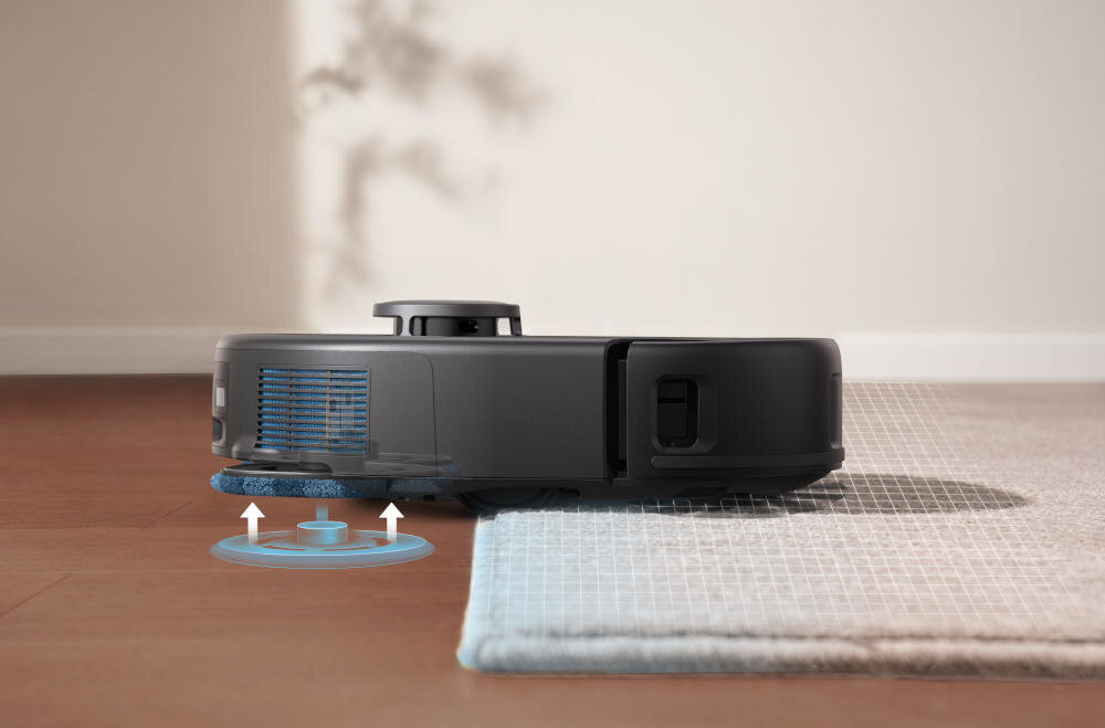 Robot sprzątający porusza się po podłodze, częściowo najeżdżając na dywan. Widać niebieskie elementy graficzne sugerujące działanie mechanizmu czyszczącego.