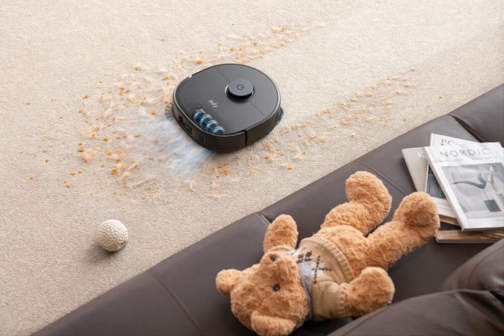 Robot sprzątający porusza się po dywanie, zbierając rozsypane okruchy. Obok leży pluszowy miś i magazyn z napisem \'NORDIC\'.