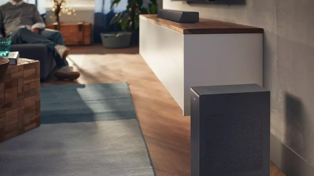 Na zdjęciu widać czarny soundbar umieszczony na drewnianej półce pod telewizorem oraz subwoofer stojący na podłodze. W tle znajduje się osoba siedząca na kanapie oraz rośliny doniczkowe.