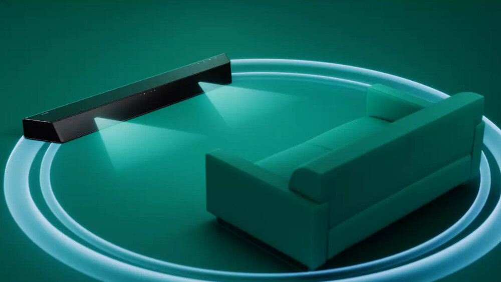 Na obrazie widoczny jest czarny soundbar ustawiony przed zieloną kanapą, z efektami świetlnymi sugerującymi dźwięk rozchodzący się wokół. Tło jest jednolite, w odcieniach zieleni.