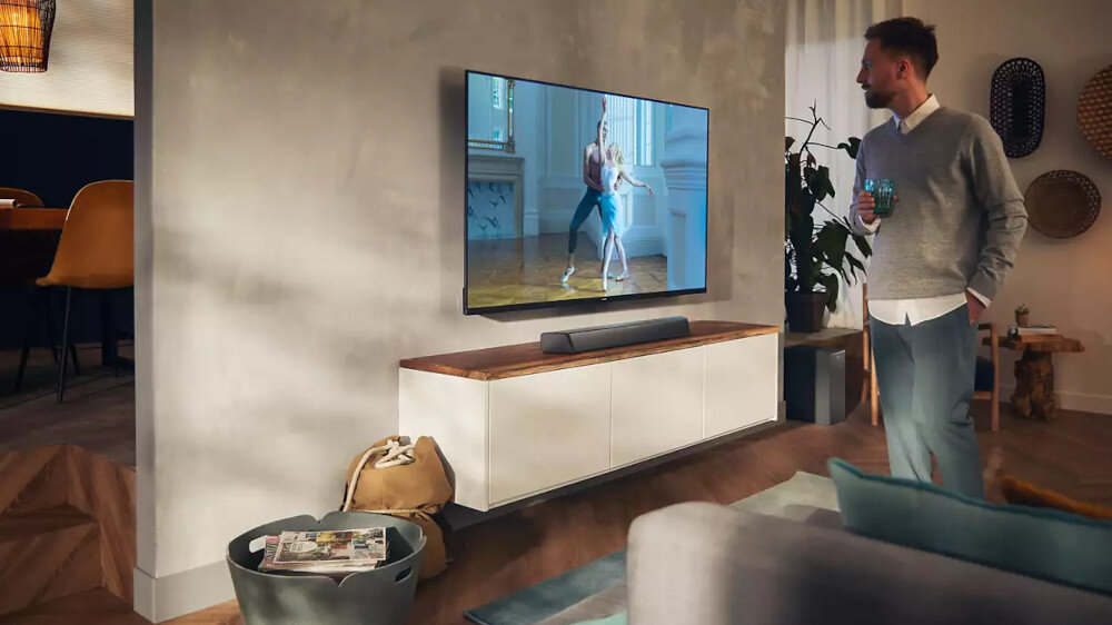 W salonie na ścianie wisi telewizor, pod którym znajduje się czarny soundbar. Obok stoi mężczyzna trzymający szklankę, a na ekranie telewizora widać osobę tańczącą balet.