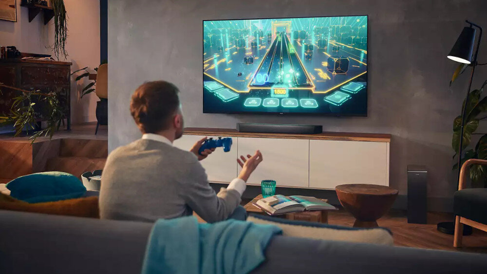Osoba siedzi na kanapie i gra na konsoli, patrząc na duży telewizor, na którym wyświetlana jest gra. Przed telewizorem znajduje się soundbar, a na stoliku leżą książki i kubek.