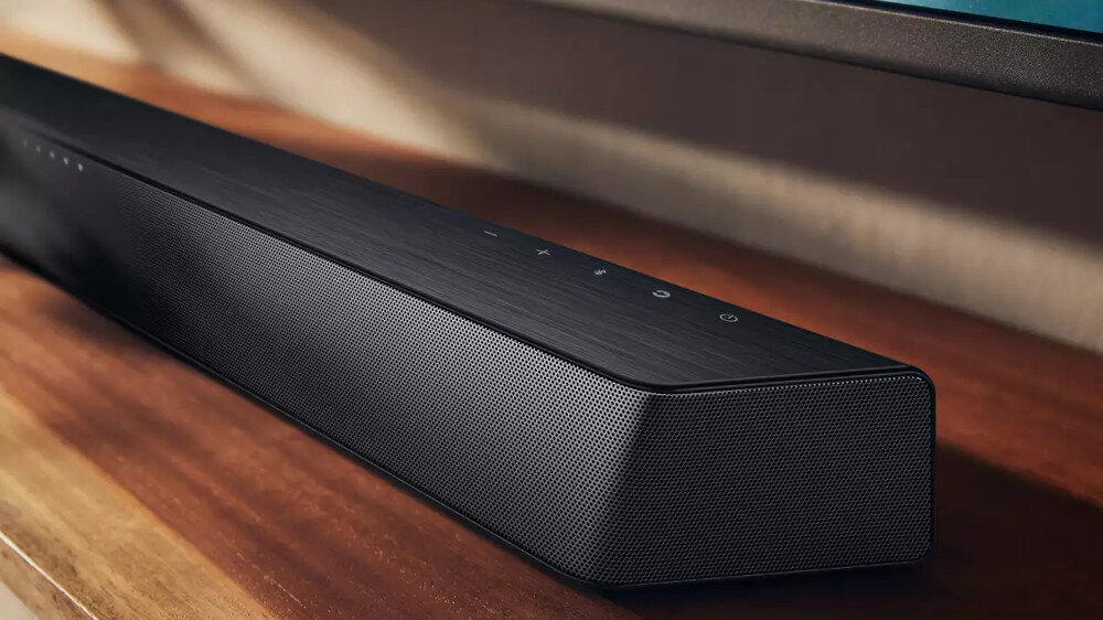 Czarny soundbar z widocznymi przyciskami dotykowymi na górnej powierzchni, umieszczony na drewnianej powierzchni. Obudowa ma metaliczną siatkę na przedniej części.